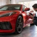 Man crouches beside a red sports car in a garage, shining a handheld light along the front fender and door panel during a paint inspection.